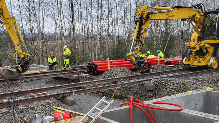 Gravemaskiner og energimontører legger rør og utfører arbeid ved jernbanespor omgitt av skog og utstyr.